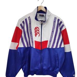 May include: A vintage zip-up track jacket in white, red, and blue. The jacket features a high collar, a red logo, and purple striped accents. The sleeves are blue and the jacket is hanging on a wooden hanger.