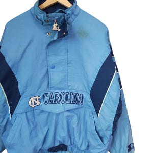May include: A light blue and navy blue Starter pullover jacket featuring the text CAROLINA and the North Carolina Tar Heels logo on the front pocket flap. The jacket has a high collar, a partial zip with a branded pull, and snap buttons.