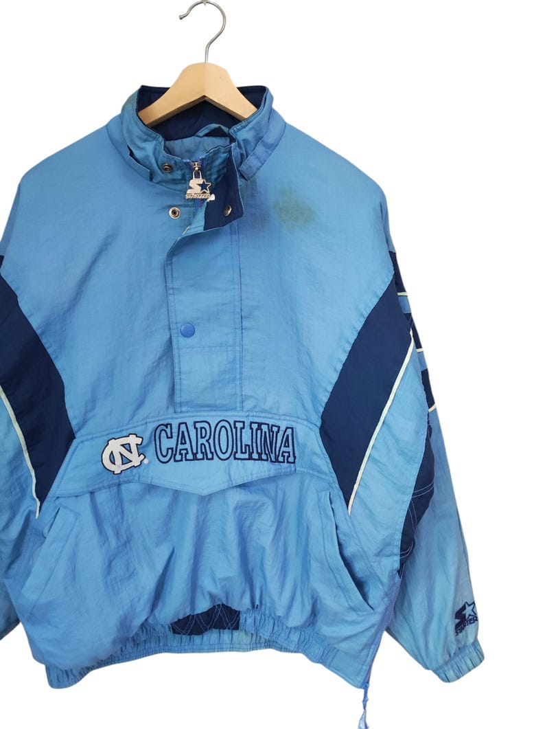 May include: A light blue and navy blue Starter pullover jacket featuring the text CAROLINA and the North Carolina Tar Heels logo on the front pocket flap. The jacket has a high collar, a partial zip with a branded pull, and snap buttons.
