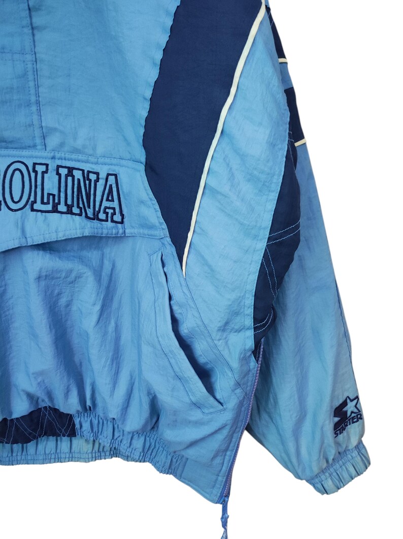 May include: A light blue and navy blue pullover windbreaker jacket featuring the text OLINA embroidered in navy blue on the front pocket flap. The jacket includes a side zip, elastic cuffs, and a Starter logo patch on the left sleeve.