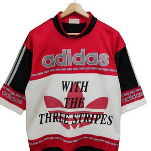 Op de afbeelding: Rood, zwart en wit Adidas T-shirt met de tekst "adidas" over de borst. Het shirt heeft het Adidas-logo en de woorden "WITH THE THREE STRIPES". De mouwen hebben het Adidas-logo.