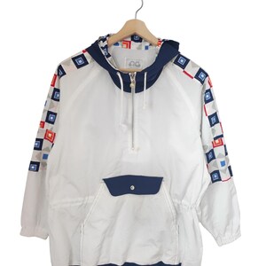 May include: White anorak jacket with a navy blue hood, pocket, and trim. The sleeves feature a geometric pattern of red, blue, and gray squares. The jacket has a half-zip closure and a drawstring waist. The jacket is hanging on a wooden hanger.