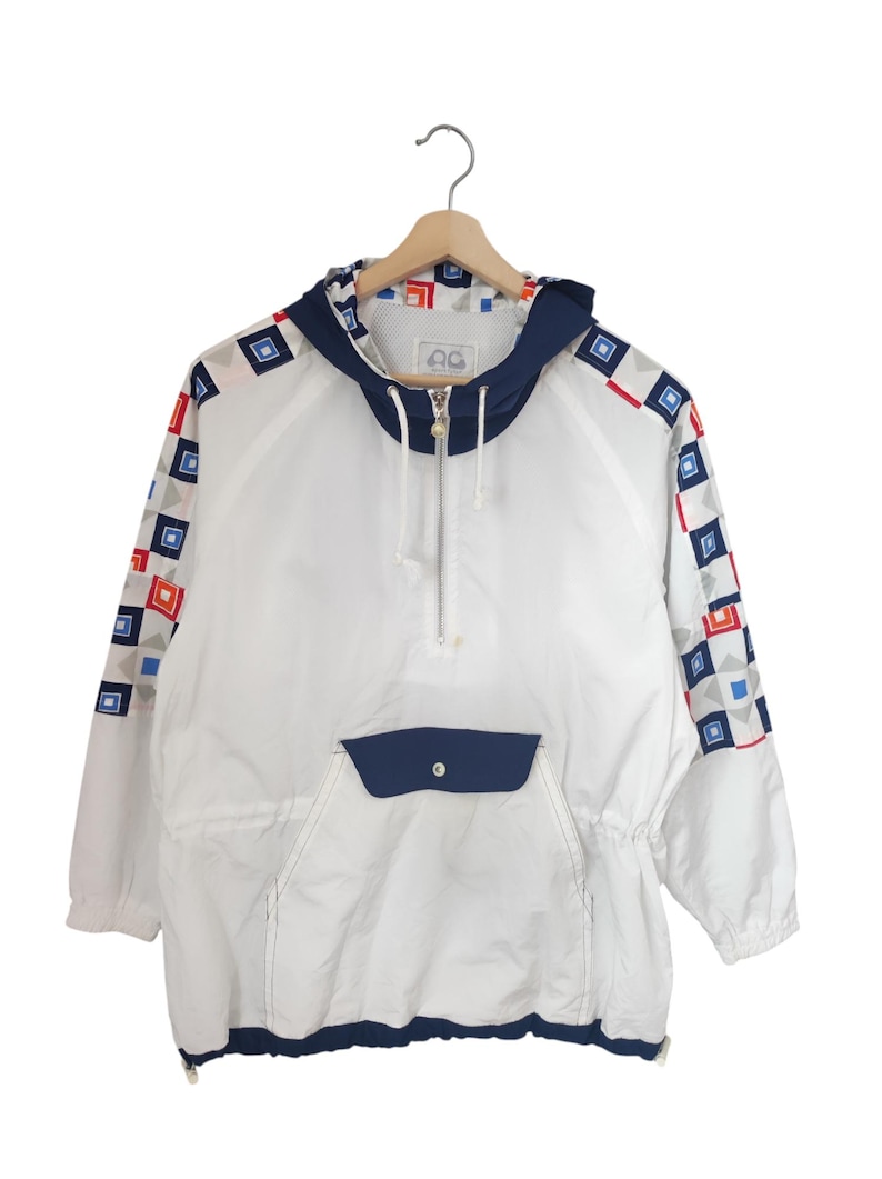 May include: White anorak jacket with a navy blue hood, pocket, and trim. The sleeves feature a geometric pattern of red, blue, and gray squares. The jacket has a half-zip closure and a drawstring waist. The jacket is hanging on a wooden hanger.