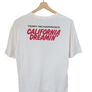 May include: White t-shirt with the text "TERRY RICHARDSON'S CALIFORNIA DREAMIN'" in red. The shirt is hanging on a wooden hanger against a white background. The t-shirt has short sleeves.