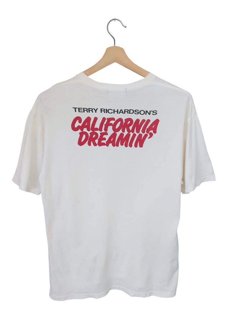 May include: White t-shirt with the text "TERRY RICHARDSON'S CALIFORNIA DREAMIN'" in red. The shirt is hanging on a wooden hanger against a white background. The t-shirt has short sleeves.