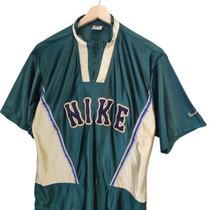 May include: Green and cream-colored Nike short-sleeve shirt with a zip-up collar. The word "NIKE" is prominently displayed across the chest in a blue and gold font. A small Nike logo is visible on the sleeve.