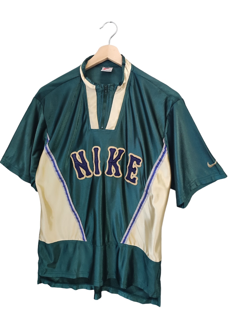 May include: Green and cream-colored Nike short-sleeve shirt with a zip-up collar. The word "NIKE" is prominently displayed across the chest in a blue and gold font. A small Nike logo is visible on the sleeve.