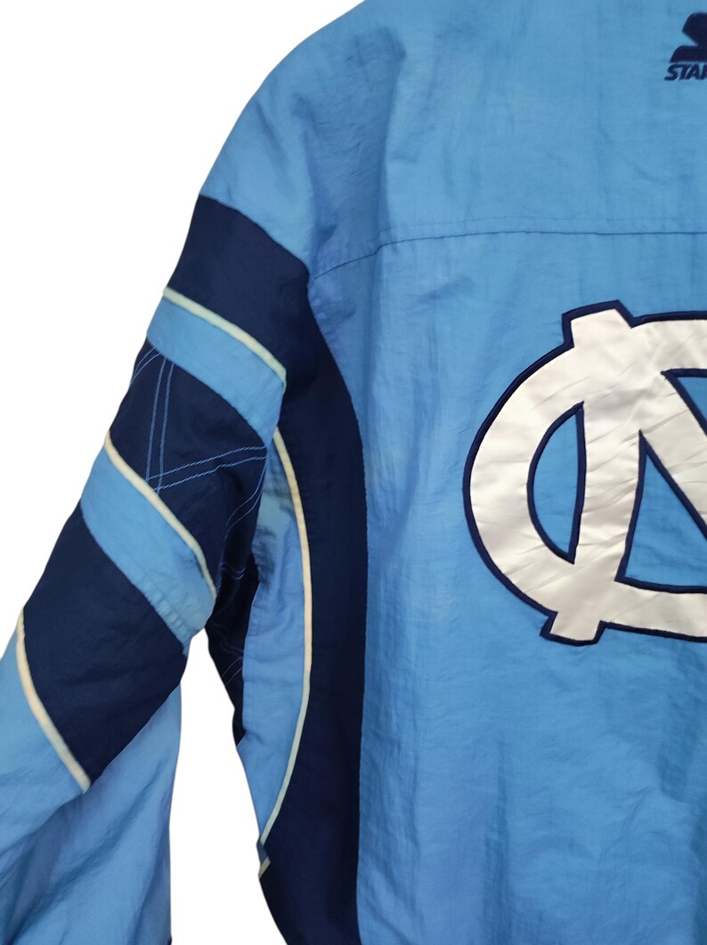 May include: A light blue and navy blue windbreaker jacket featuring a large white embroidered NC logo on the back. The jacket includes a Starter brand logo near the collar, long sleeves with navy blue and light blue stripes, and white piping.