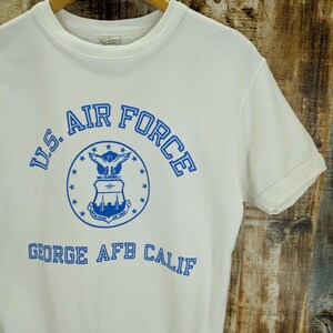 May include: Cream-coloured short-sleeved sweatshirt featuring the text "U.S. AIR FORCE" in blue, a blue emblem, and the text "GEORGE AFB CALIF". The sweatshirt is displayed on a wooden hanger against a wood-grain backdrop.