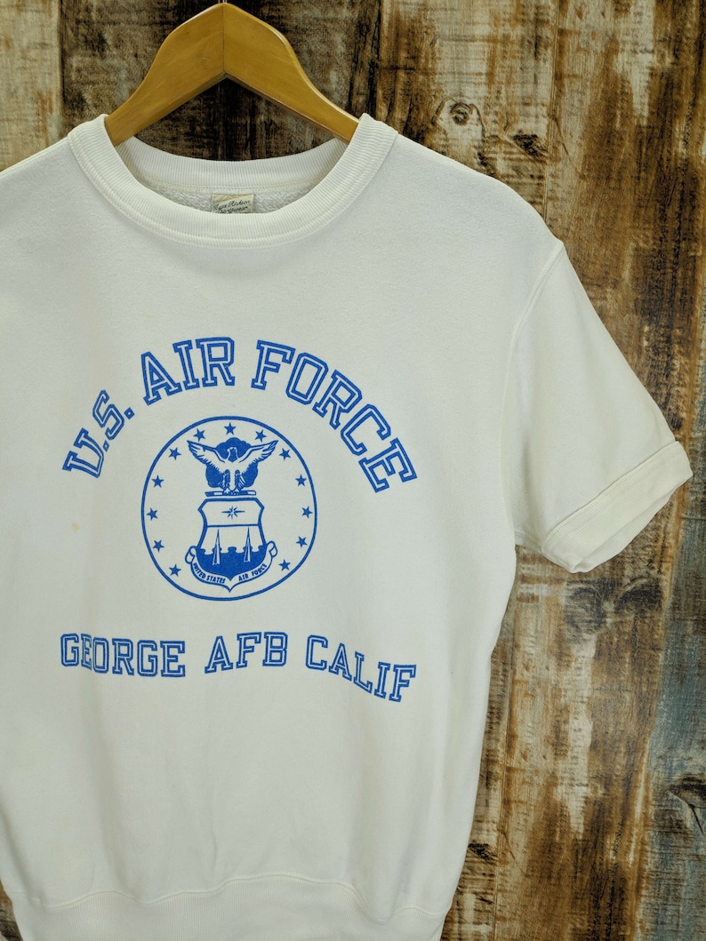 May include: Cream-coloured short-sleeved sweatshirt featuring the text "U.S. AIR FORCE" in blue, a blue emblem, and the text "GEORGE AFB CALIF". The sweatshirt is displayed on a wooden hanger against a wood-grain backdrop.
