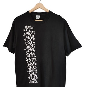 May include: Black t-shirt with a white graphic of stylized figures arranged vertically. The design is a repeating pattern of stick-figure-like people. The shirt is displayed on a wooden hanger.