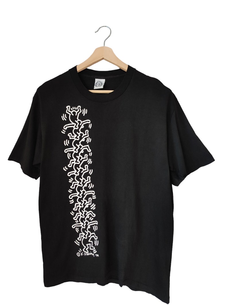 May include: Black t-shirt with a white graphic of stylized figures arranged vertically. The design is a repeating pattern of stick-figure-like people. The shirt is displayed on a wooden hanger.