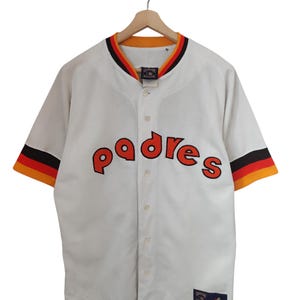 May include: White baseball jersey with the word "padres" in orange lettering across the chest. The jersey has orange, black, and yellow trim on the sleeves and collar. The jersey is button-down and hangs on a wooden hanger.