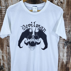 May include: White t-shirt featuring a black graphic of a skull with horns and the word "Devilman" in a gothic font. The shirt is displayed on a wooden hanger against a wood-grain background.