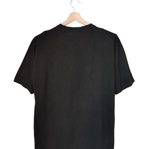 May include: A plain black t-shirt is suspended from a wooden hanger against a white backdrop. The short-sleeved shirt is made of a soft material and is viewed from the back, highlighting its simple design. The hanger is made of light-coloured wood.