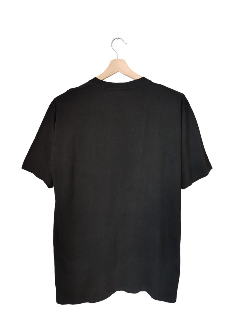 May include: A plain black t-shirt is suspended from a wooden hanger against a white backdrop. The short-sleeved shirt is made of a soft material and is viewed from the back, highlighting its simple design. The hanger is made of light-coloured wood.