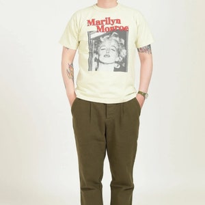 May include: A person wearing a light yellow t-shirt with a black and white image of Marilyn Monroe and the text "Marilyn Monroe". The person is also wearing olive green pants, a blue hat, sunglasses, and black sneakers.