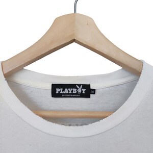 May include: Close-up of a white t-shirt with a crew neck, hanging on a wooden hanger. The shirt features a black label with the Playboy logo and the text "PLAYBOY HYSTERIC GLAMOUR". The shirt is a size M.