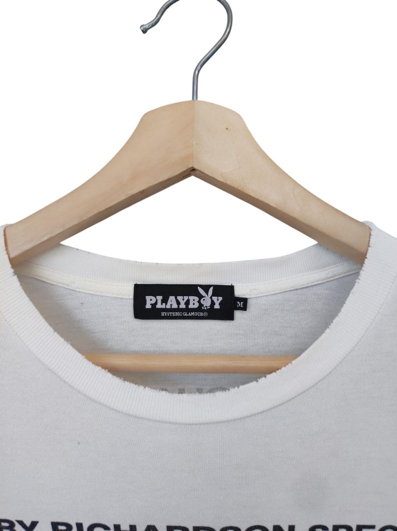 May include: Close-up of a white t-shirt with a crew neck, hanging on a wooden hanger. The shirt features a black label with the Playboy logo and the text "PLAYBOY HYSTERIC GLAMOUR". The shirt is a size M.
