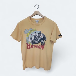 May include: Mustard yellow t-shirt featuring a Batman graphic. The graphic shows Batman and another figure on a motorcycle with the word "WROOM!" and the word "BATMAN" in red. The shirt is hanging on a wooden hanger.