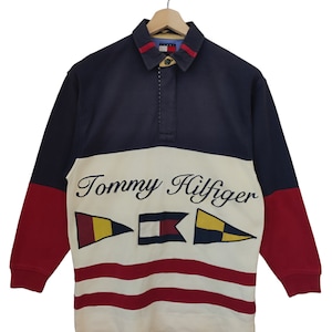 May include: A long-sleeved Tommy Hilfiger rugby shirt. The shirt features a navy blue collar and sleeves, a white midsection with the brand name and flag graphics, and red sleeves. The shirt is hanging on a wooden hanger.