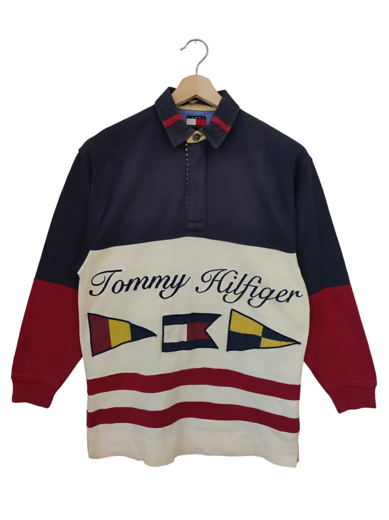May include: A long-sleeved Tommy Hilfiger rugby shirt. The shirt features a navy blue collar and sleeves, a white midsection with the brand name and flag graphics, and red sleeves. The shirt is hanging on a wooden hanger.
