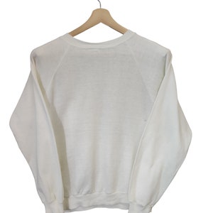 May include: A white crew neck sweatshirt hanging on a wooden hanger. The long-sleeved top has a ribbed hem and cuffs. The raglan sleeves extend from the neckline to the underarm. The sweatshirt is made of a soft, lightweight material.