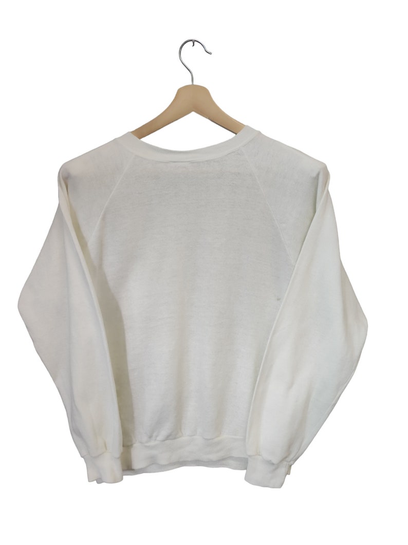 May include: A white crew neck sweatshirt hanging on a wooden hanger. The long-sleeved top has a ribbed hem and cuffs. The raglan sleeves extend from the neckline to the underarm. The sweatshirt is made of a soft, lightweight material.