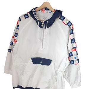 May include: White hooded anorak jacket with a navy blue hood and pocket. The sleeves feature a geometric pattern of red, blue, and gray squares. The jacket has a half-zip closure and a drawstring at the waist.