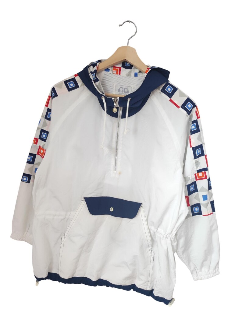 May include: White hooded anorak jacket with a navy blue hood and pocket. The sleeves feature a geometric pattern of red, blue, and gray squares. The jacket has a half-zip closure and a drawstring at the waist.