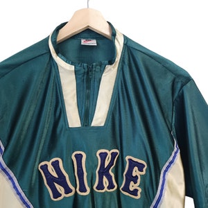 May include: Teal and cream-colored vintage Nike pullover shirt with a quarter-zip closure. The shirt features the Nike logo in a navy blue, varsity-style font. The shirt is hanging on a wooden hanger against a white background.