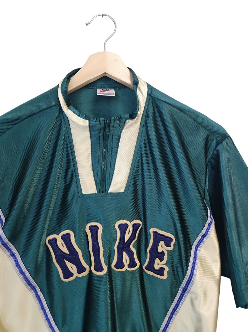 May include: Teal and cream-colored vintage Nike pullover shirt with a quarter-zip closure. The shirt features the Nike logo in a navy blue, varsity-style font. The shirt is hanging on a wooden hanger against a white background.
