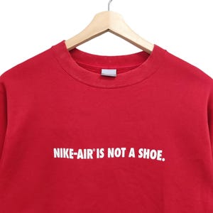 May include: A red crew neck sweatshirt with the text "NIKE-AIR IS NOT A SHOE." in white. The sweatshirt is hanging on a wooden hanger against a white background.
