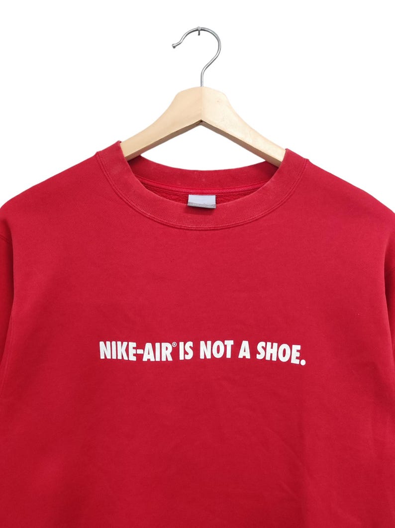 May include: A red crew neck sweatshirt with the text "NIKE-AIR IS NOT A SHOE." in white. The sweatshirt is hanging on a wooden hanger against a white background.