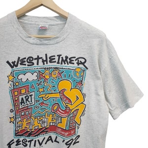 May include: A light gray t-shirt featuring a colorful graphic for the Westheimer Art Festival '92. The design includes a yellow figure playing a saxophone, buildings, and the word "ART". The shirt is hanging on a wooden hanger.