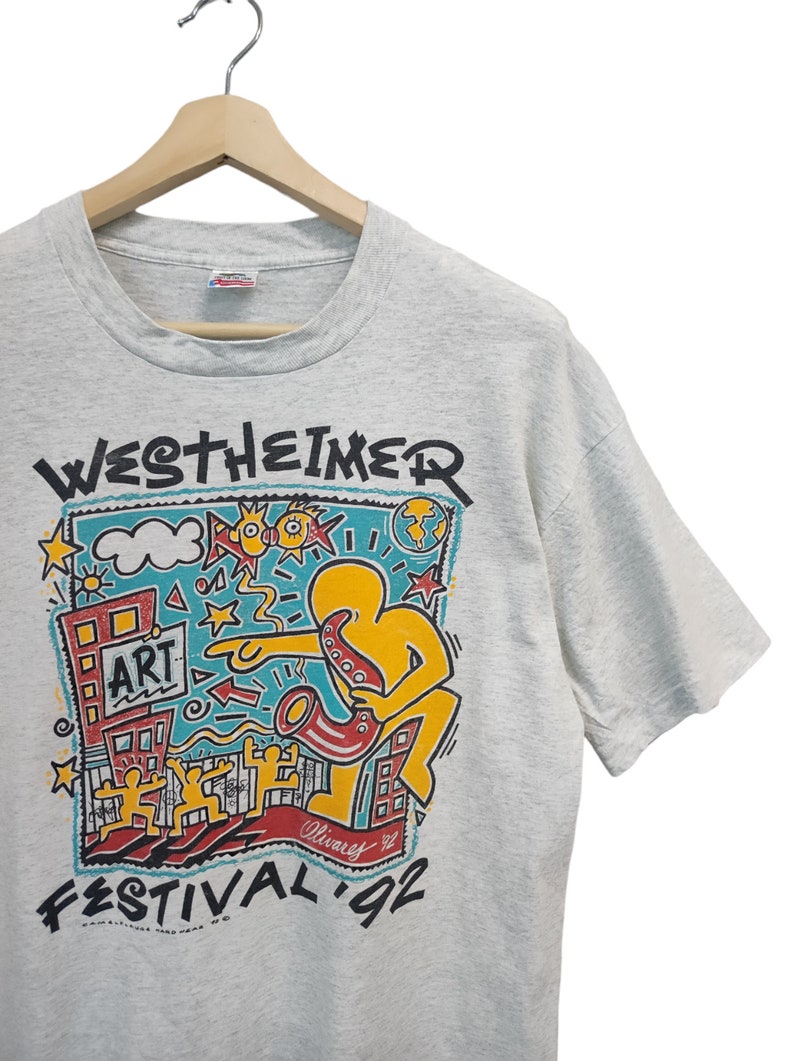 May include: A light gray t-shirt featuring a colorful graphic for the Westheimer Art Festival '92. The design includes a yellow figure playing a saxophone, buildings, and the word "ART". The shirt is hanging on a wooden hanger.