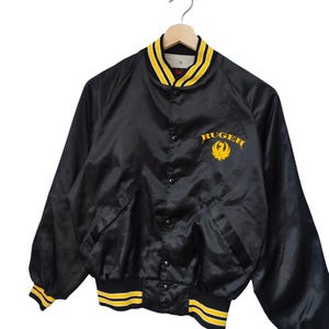 May include: Black satin bomber jacket with yellow and white striped trim. The jacket features the word "Ruger" in yellow script on the chest, along with a yellow emblem. The collar and cuffs have yellow and white stripes.