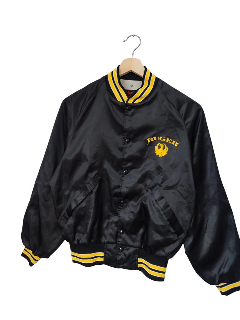 May include: Black satin bomber jacket with yellow and white striped trim. The jacket features the word "Ruger" in yellow script on the chest, along with a yellow emblem. The collar and cuffs have yellow and white stripes.