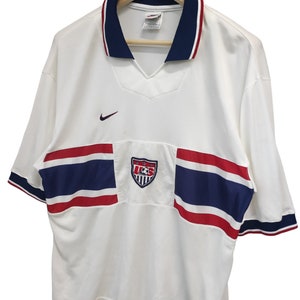 May include: White football shirt with blue and red stripes and trim. Includes a U.S. Soccer crest and Nike logo. Short-sleeved with a v-neck collar. The shirt is on a wooden hanger.