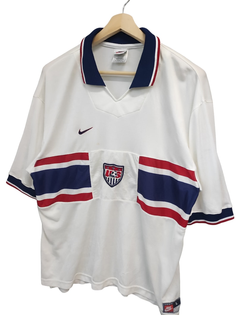 May include: White football shirt with blue and red stripes and trim. Includes a U.S. Soccer crest and Nike logo. Short-sleeved with a v-neck collar. The shirt is on a wooden hanger.