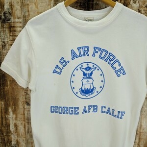 May include: A white short-sleeved sweatshirt with the text "U.S. AIR FORCE" in blue, encircling a blue emblem. Below the emblem is the text "GEORGE AFB CALIF" in blue. The sweatshirt is hanging on a wooden hanger.