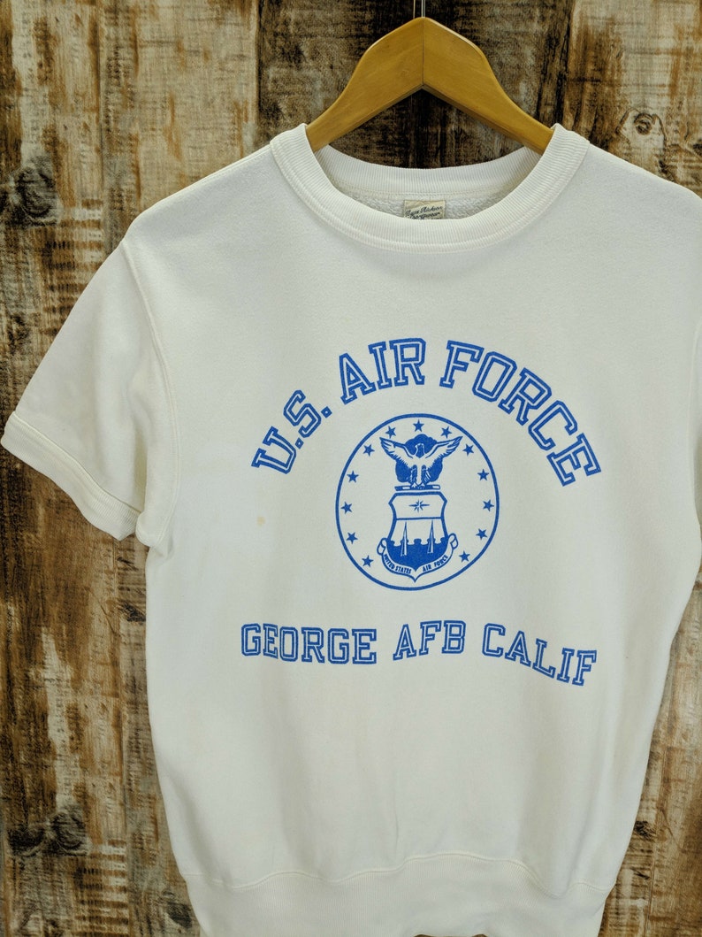 May include: A white short-sleeved sweatshirt with the text "U.S. AIR FORCE" in blue, encircling a blue emblem. Below the emblem is the text "GEORGE AFB CALIF" in blue. The sweatshirt is hanging on a wooden hanger.