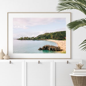 May include: A framed photograph of a beach scene with a rocky outcropping in the foreground. The beach is sandy and there are people swimming and sunbathing. The water is a clear turquoise color and the sky is a light blue with white clouds.