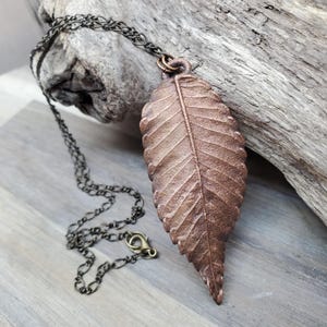 May include: A bronze-colored leaf pendant necklace. The pendant is a detailed replica of a leaf, with visible veins and texture. The necklace chain is dark, with a lobster clasp. The pendant is approximately 3 inches long.