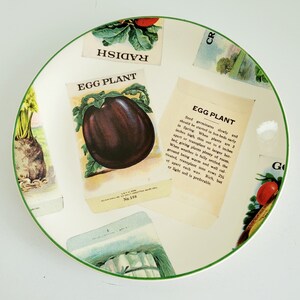 Könnte beinhalten: Ein weißer Keramikteller mit grünem Rand, verziert mit Vintage-Saatgutpackungs-Illustrationen. Das zentrale Bild ist eine Aubergine, mit dem Text "EGG PLANT" und Pflanzanweisungen. Andere Packungen zeigen Radieschen und anderes Gemüse.