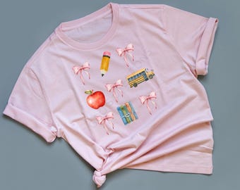 Puede incluir: Camiseta rosa claro con ilustraciones de elementos escolares. El diseño incluye una manzana roja, un lápiz amarillo, un autobús escolar amarillo, libros y lazos rosas.