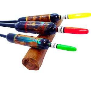 May include: Three decorative fishing floats with black bodies, colourful tips, and painted designs. The tips are yellow, red, and green. The floats rest on a wooden support, showcasing their unique craftsmanship.