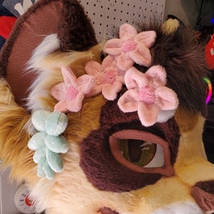 May include: A light brown and white furry animal head with a brown nose and green and pink flower accents. The head is a furry costume.