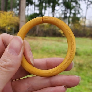 Puede incluir: Una pulsera de madera de color amarillo mostaza con un diseño tallado y texturizado. La pulsera se sostiene en la mano de una persona sobre un fondo de hierba verde y árboles. La pulsera es un círculo completo, con un pequeño espacio.