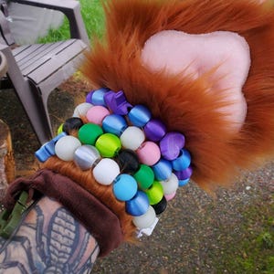 Könnte beinhalten: Ein pelziger, pfotenförmiger Handschuh mit einer braunen Manschette und einem Perlenarmband. Die Pfote hat braunes Fell, olivgrüne Ballen und weiße Krallen. Das Armband hat verschiedenfarbige Perlen, darunter blau, grün, lila und weiß.